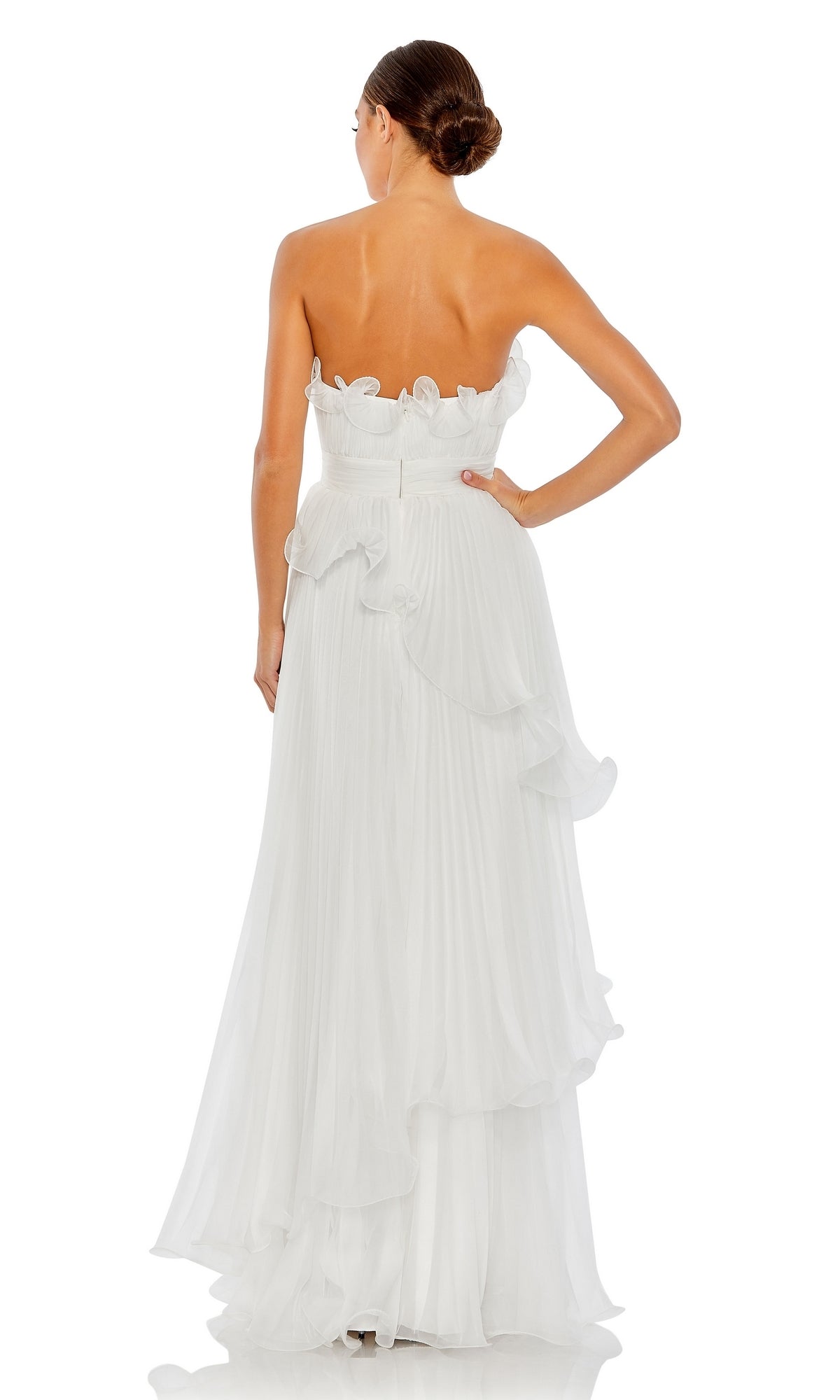 A woman stands facing away, wearing the Long Formal Dress 49537 by Mac Duggal—a strapless white gown with ruffles, a pleated floor-length skirt, and a subtle empire waist. Her hair is styled in a low bun.